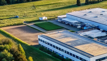 Aerial view of a white, flat-roofed industrial building with parking and green fields surrounding it, bordered by trees and hedges.