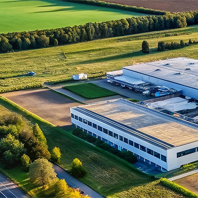 Aerial view of a white, flat-roofed industrial building with parking and green fields surrounding it, bordered by trees and hedges.