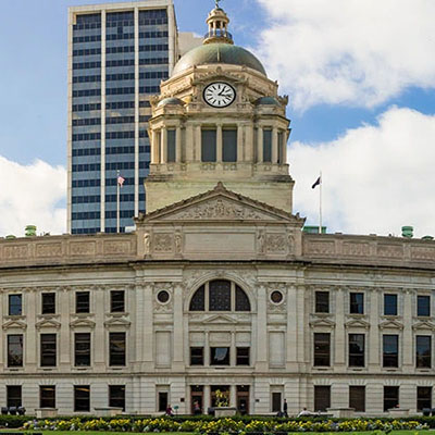 Front view of Fort Wayne courthouse with a clock tower and dome, with a modern skyscraper behind and a blue sky above